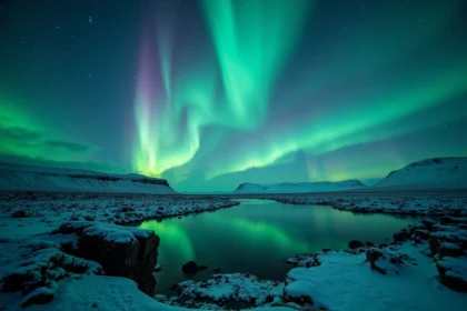 Aurore boréale verte et violette reflétée dans un lac glaciaire en Islande avec des roches basaltiques enneigées au premier plan