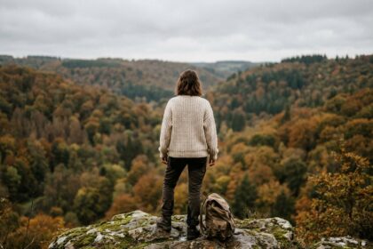 Femme en pull chunky observant la vallée boisée de l'Ardenne en automne depuis un rocher moussu